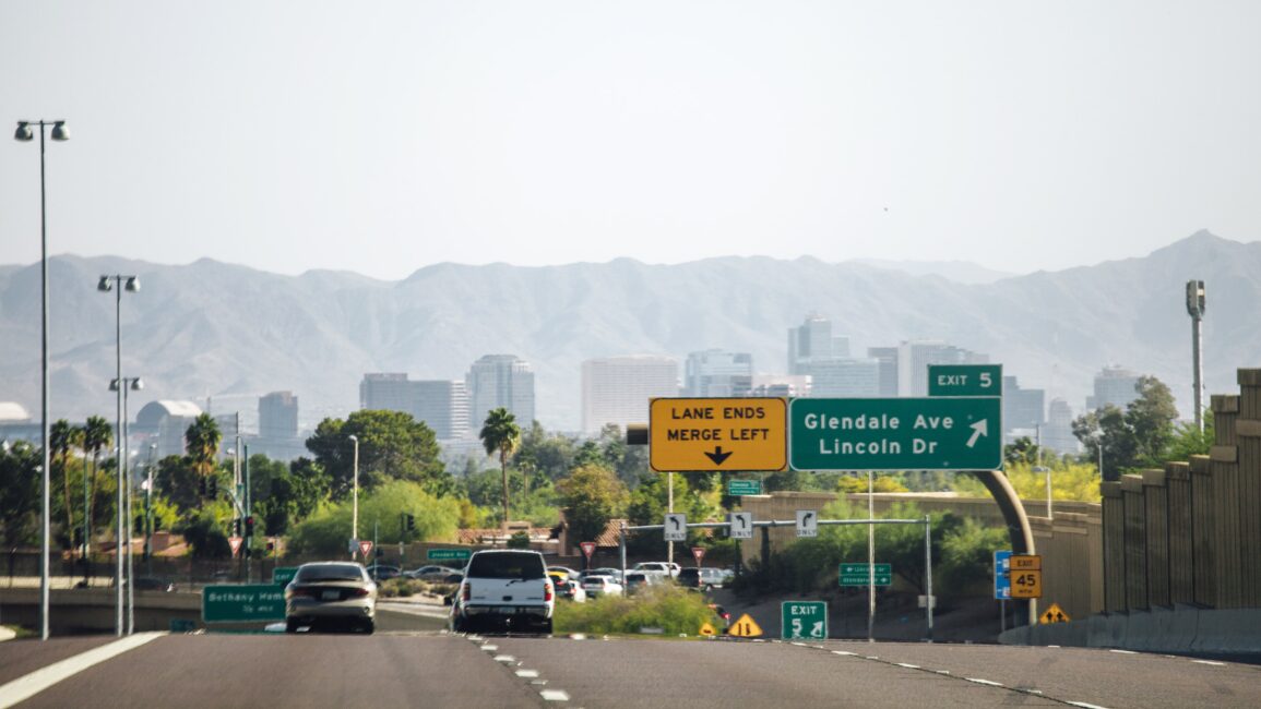 Highway scene with vehicles driving towards a city skyline and mountains in the background. Signs indicate exit 5 for Glendale Ave and Lincoln Dr, and a lane ending with a merge left instruction, raising the question of who gets the insurance check when a car is totaled in such moments.